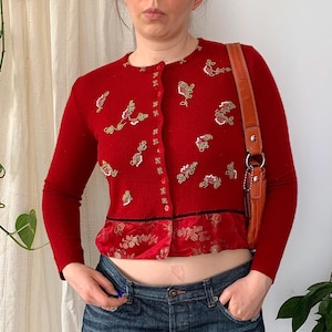 Puede incluir: Un cárdigan rojo con bordados florales y un panel de satén, combinado con una minifalda vaquera. El cárdigan tiene mangas largas y botones. Un bolso de hombro de cuero marrón completa el conjunto.