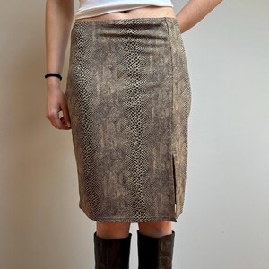 Peut inclure: Jupe mi-longue à motif peau de serpent dans les tons marron et beige. La jupe a une fente sur le côté. Portée avec de hautes bottes en cuir marron foncé.