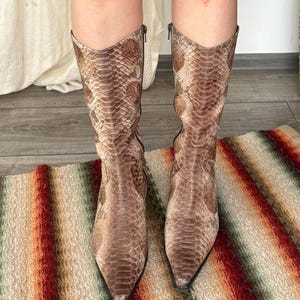 May include: A pair of mid-calf boots featuring a brown and tan snakeskin pattern. The boots have a pointed toe, a V-shaped topline, and a vertical zip closure on the inner side of each shaft, extending from the top down to the ankle area.