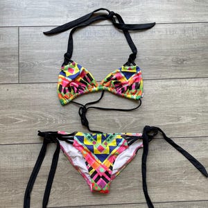 Puede incluir: Un conjunto de bikini de dos piezas con un vibrante patrón geométrico multicolor en tonos rosa, amarillo, azul y verde. La parte superior triangular y la braga de bikini con lazos laterales incluyen tiras negras lisas para ajustar.
