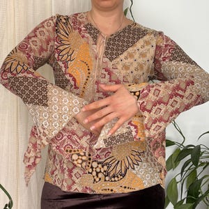 May include: A long-sleeved blouse featuring a patchwork pattern in shades of brown, tan, orange, and cream with a lace-up neckline and flared sleeves, paired with a dark brown velvet skirt. The blouse displays various floral and geometric motifs.