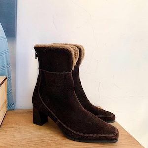 May include: Brown suede ankle boots with a square toe and block heel. The boots feature a faux fur lining and a rear zip. The boots are displayed on a wooden surface. The heel height is approximately 7 cm.