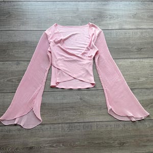May include: A light pink long-sleeve top featuring a wrap-style front with a V-neckline and wide, flared sleeves. The sleeves are constructed with a pleated texture that extends from the shoulder to the scalloped, open-ended cuffs.