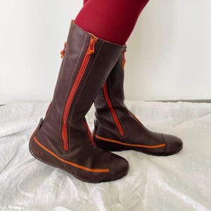 May include: Brown leather knee-high boots with bright orange zippers and accents. The boots have a rounded toe and a flat sole. The zippers run down the front and back of the boots. The boots are worn with red leggings.