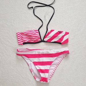 May include: A two-piece swimsuit in white and hot pink stripes. The top is a bandeau style with a halter neck, and the bottoms are a classic bikini cut. The swimsuit has a black trim and a small logo on the top.