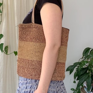 Puede incluir: Un bolso tote tejido con forma rectangular. El bolso presenta rayas horizontales en tonos marrones y beige. Tiene dos correas beige y un cierre de cremallera. El bolso se lleva al hombro.