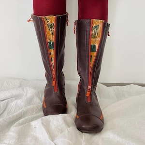 May include: A pair of knee-high dark brown boots featuring a front-facing orange zipper closure. The boots include a vertical panel of patterned fabric in shades of green, yellow, and brown running alongside the zipper, with orange trim accents.