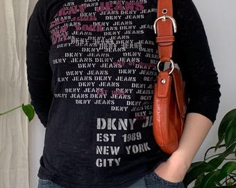 Op de afbeelding: Zwart shirt met lange mouwen met roze en witte tekst "DKNY JEANS". De outfit omvat een blauwe jeans en een bruine leren schoudertas met een zilveren gesp. De tasriem is ongeveer 2,5 cm breed.