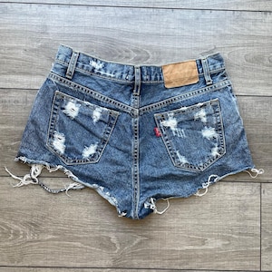 May include: Distressed denim shorts with a frayed hem and a back pocket with a red label. The shorts are a light blue wash and have a relaxed fit.