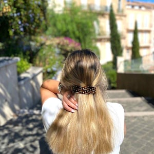 Könnte beinhalten: Ein brauner und schwarzer rechteckiger Haarclip hält blondes Haar zurück. Der Clip hat ein gewebtes Design und ist horizontal positioniert. Die Person trägt ein weißes Oberteil und eine silberne Armbanduhr.