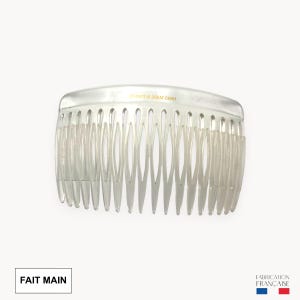 Pode incluir: Um pente de cabelo de plástico transparente com dentes finos. O pente tem um topo curvo e está rotulado "HAND MADE IN FRANCE" em letras douradas. As palavras "FAIT MAIN" estão em uma caixa branca no canto inferior esquerdo.