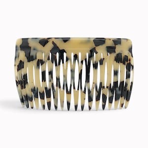 Pode incluir: Um pente de cabelo com uma borda superior retangular e uma série de dentes longos e afilados. O acessório é feito de um material liso e polido com um padrão manchado de creme, preto e castanho escuro em toda a sua superfície.
