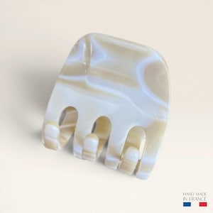 Peut inclure: Une pince à cheveux carrée avec un motif marbré translucide dans des tons crème, beige et blanc. L'accessoire possède quatre dents arrondies et comporte le texte "HAND MADE IN FRANCE" dans le coin inférieur droit.