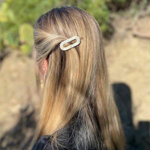 May include: A gold-toned hair clip in an oval shape, adorned with a marbled pattern, is clipped into blonde hair. The clip is positioned on the side of the head, adding a decorative touch to the hairstyle. The background is blurred, with hints of greenery.