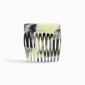 Könnte beinhalten: Ein Haarkamm mit einer rechteckigen Oberkante und elf nach unten gerichteten Zinken. Das Material weist ein marmoriertes Muster mit Streifen in Creme, Beige und Dunkelanthrazit auf, was eine geschichtete, lineare Optik auf der Oberfläche erzeugt.