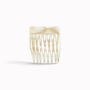 Könnte beinhalten: Ein Haarkamm mit einer glatten, polierten Oberfläche in einem marmorierten Creme- und Off-White-Muster. Das Accessoire hat eine rechteckige Oberkante und acht spitz zulaufende, gebogene Zinken, die dazu dienen, das Haar sicher zu halten.