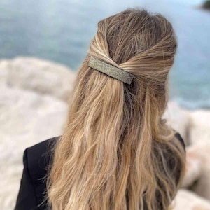 May include: A rectangular hair clip with a glittery, textured surface is fastened into blonde hair. The clip is a dark colour, possibly black or dark grey, and contrasts with the lighter hair. The hair is styled in a half-up, half-down style.