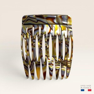 Könnte beinhalten: Ein Haarkamm mit einem Marmormuster in Braun-, Gold- und Grautönen mit glänzender Oberfläche. Der Kamm hat einen breiten, geschwungenen oberen Rand und neun spitz zulaufende Zinken. Der Text "HAND MADE IN FRANCE" steht in der unteren Ecke.