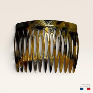 May include: A hair comb featuring a tortoiseshell pattern with dark brown and amber tones. The comb has a curved top edge and eleven long, pointed teeth with oval-shaped cutouts between them. The text HAND MADE IN FRANCE is printed at the bottom.