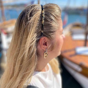 May include: A tortoiseshell patterned headband worn by a person with blonde hair. The headband is a thin, rectangular shape with a repeating pattern of dark brown and cream colours. The person is also wearing gold-toned earrings.