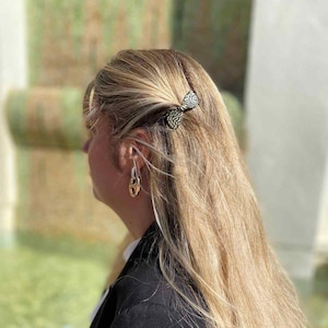Peut inclure: Un nœud papillon noir et blanc est fixé dans des cheveux blonds. Des boucles d'oreilles dorées et argentées sont visibles. La personne porte un blazer noir. Accessoire de coiffure décoratif.