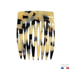 Könnte beinhalten: Ein Haarkamm mit Schildpattmuster mit gewölbter Oberseite und mehreren Zähnen. Der Kamm hat eine Mischung aus schwarzen, braunen und cremefarbenen Farben. Die Worte "HAND MADE IN FRANCE" sind unten rechts aufgedruckt.