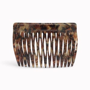 May include: A hair comb featuring a tortoiseshell pattern with a blend of dark brown, light brown, and translucent amber tones. The accessory has a smooth, curved top edge and a series of fourteen long, tapered teeth designed for styling hair.