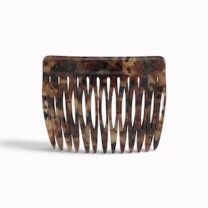Könnte beinhalten: Ein Haarkamm mit einer geschwungenen Oberkante und vierzehn langen, spitz zulaufenden Zinken. Das Accessoire zeigt ein gesprenkeltes Schildpattmuster in Dunkelbraun-, Bernstein- und Beigetönen mit einer glatten, polierten Oberfläche.