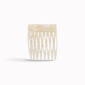 May include: A hair comb featuring a rectangular top edge and seven tapered teeth. The accessory is crafted from a smooth, semi-translucent material with a cream and off-white marbled pattern that creates a subtle, light-reflecting appearance.