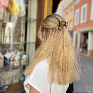 Peut inclure: Une pince à cheveux blonde à motif écaille de tortue retient une partie de longs cheveux blonds raides. La personne porte un haut blanc. L'arrière-plan montre une devanture de magasin et des bâtiments.