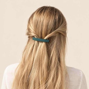 Op de afbeelding: Een rechthoekige haarclip in een effen donkergroenblauwe kleur die een sectie lang blond haar vastzet. Het haar is gestyled in een half-opgestoken kapsel waarbij twee zijlokken zijn gedraaid en naar het midden van het hoofd zijn gebracht.