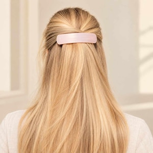 Op de afbeelding: Een rechthoekige haarspeld in een zachte, lichtroze tint met een gladde, matte afwerking. Het haaraccessoire is horizontaal geplaatst en houdt een sectie van lang, steil blond haar aan de achterkant van het hoofd vast.