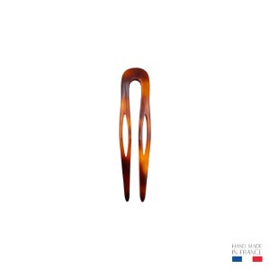 Puede incluir: Una horquilla para el cabello con un patrón de carey con una parte superior en forma de U y dos púas. El accesorio para el cabello tiene un recorte ovalado en cada púa. Las palabras "HAND MADE IN FRANCE" están impresas en la parte inferior derecha.