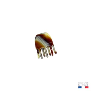 Peut inclure: Une petite pince à cheveux marron, blanche et beige. La pince a un dessus incurvé avec un motif rayé et plusieurs dents orientées vers le bas. Les mots "HAND MADE IN FRANCE" sont imprimés en bas à droite.