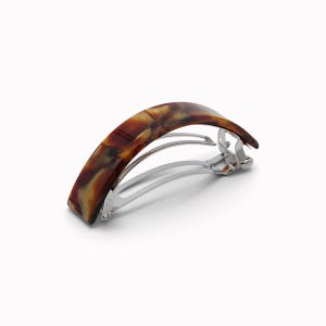 Op de afbeelding: Een gebogen haarklem met een gepolijst schildpadpatroon in tinten bruin, amber en zwart. Het accessoire is voorzien van een zilverkleurig metalen veermechanisme dat is ontworpen om het haar op zijn plaats te houden met een scharniersluiting.
