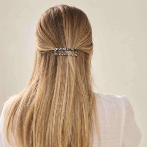 Könnte beinhalten: Eine rechteckige Haarspange mit einem Marmormuster in Blau-, Schwarz- und Weißtönen. Die Spange ist horizontal angebracht und hält eine Partie langes, glattes, hellblondes Haar. Das Accessoire hat ein Design mit einer hohlen Mitte.