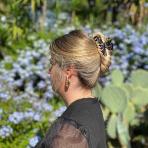 Può includere: Acconciatura bionda fissata con una clip per capelli tartarugata nera e marrone. La persona indossa orecchini a cerchio dorati e un top nero. Lo sfondo presenta piante verdi e fiori blu.