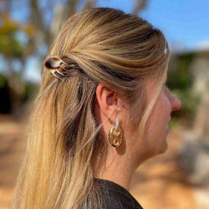 Peut inclure: Une pince à cheveux en écaille de tortue maintient les cheveux blonds. Des boucles d'oreilles dorées avec un motif de maillons de chaîne et de petites pierres blanches sont visibles. L'accessoire capillaire est un ajout élégant à la coiffure.