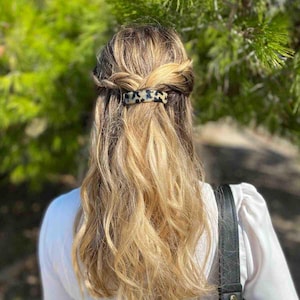 May include: A woman's blonde and brown hair is styled with a half-up, half-down braided hairstyle. A tortoiseshell patterned hair clip secures the braid. The background is a blurred green, suggesting an outdoor setting.