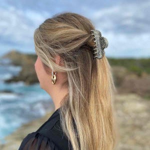 Peut inclure: Une pince à cheveux à motif noir et argenté retient des cheveux blonds. La pince est un accessoire décoratif. Des boucles d'oreilles créoles dorées sont visibles. L'arrière-plan montre une vue floue de la mer et du ciel.