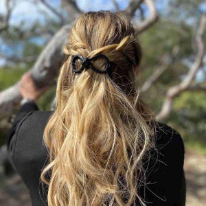 Peut inclure: Une pince à cheveux noire en forme de nœud maintient une partie de longs cheveux blonds ondulés. Les cheveux sont coiffés en demi-queue, avec la pince placée à l'arrière de la tête. L'arrière-plan est flou et verdoyant.