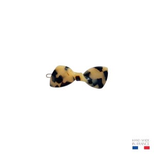 Può includere: Un fermaglio per capelli a forma di fiocco con un motivo tartarugato color crema e marrone scuro. La clip è realizzata in materiale lucido e ha una chiusura in metallo color oro. Il testo "HAND MADE IN FRANCE" è in basso a destra.
