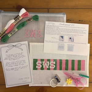 May include: A needlepoint kit featuring a mesh pouch, a canvas with a green and pink striped pattern and the letters SWS, three skeins of embroidery floss in white, pink, and green, a metal keychain ring, scissors, a needle, and instructional cards.