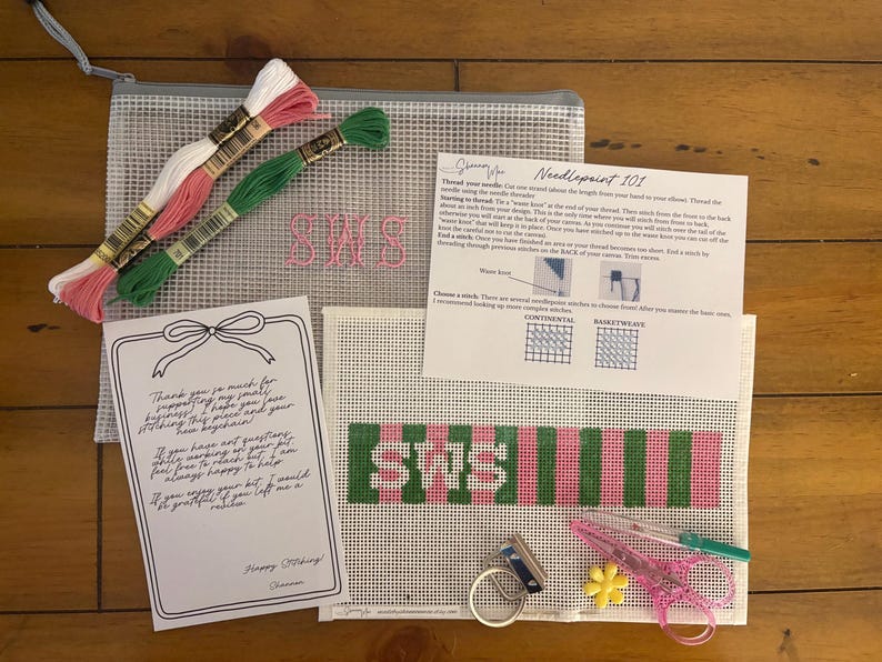 May include: A needlepoint kit featuring a mesh pouch, a canvas with a green and pink striped pattern and the letters SWS, three skeins of embroidery floss in white, pink, and green, a metal keychain ring, scissors, a needle, and instructional cards.