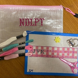 May include: A clear mesh zip-up pouch with pink trim and the text NDLPT, a blue-bordered needlepoint canvas featuring a pink plaid pattern with the letters JVK and SM, four embroidery floss skeins, a yellow flower bead, and pink scissors.