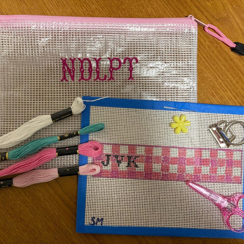 May include: A clear mesh zip-up pouch with pink trim and the text NDLPT, a blue-bordered needlepoint canvas featuring a pink plaid pattern with the letters JVK and SM, four embroidery floss skeins, a yellow flower bead, and pink scissors.