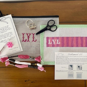 May include: A needlepoint kit with a canvas featuring the letters "Lyl" in pink and white stripes. The kit includes embroidery floss, scissors, a keychain, and a card with a handwritten message. A pink flower and pen are also included.