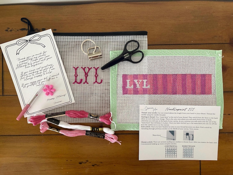 May include: A needlepoint craft kit featuring a clear mesh pouch, a canvas with a pink and purple striped pattern and the letters LYL, a metal keychain finding, black scissors, a pink needle threader, embroidery floss, and two instruction cards.
