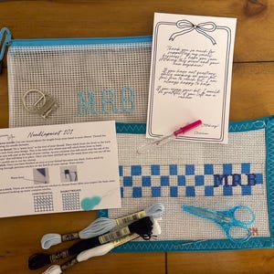 May include: A needlepoint kit featuring a mesh pouch with the letters MRB, a canvas with a blue and white checkered pattern and MRB monogram, embroidery floss in blue, black, and white, a metal keychain, a needle threader, scissors, and instructional cards.