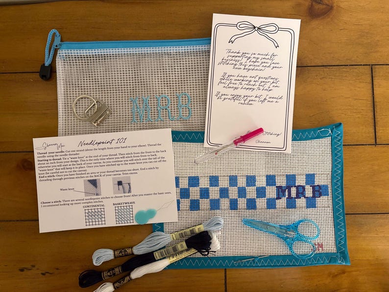 May include: A needlepoint kit featuring a mesh pouch with the letters MRB, a canvas with a blue and white checkered pattern and MRB monogram, embroidery floss in blue, black, and white, a metal keychain, a needle threader, scissors, and instructional cards.